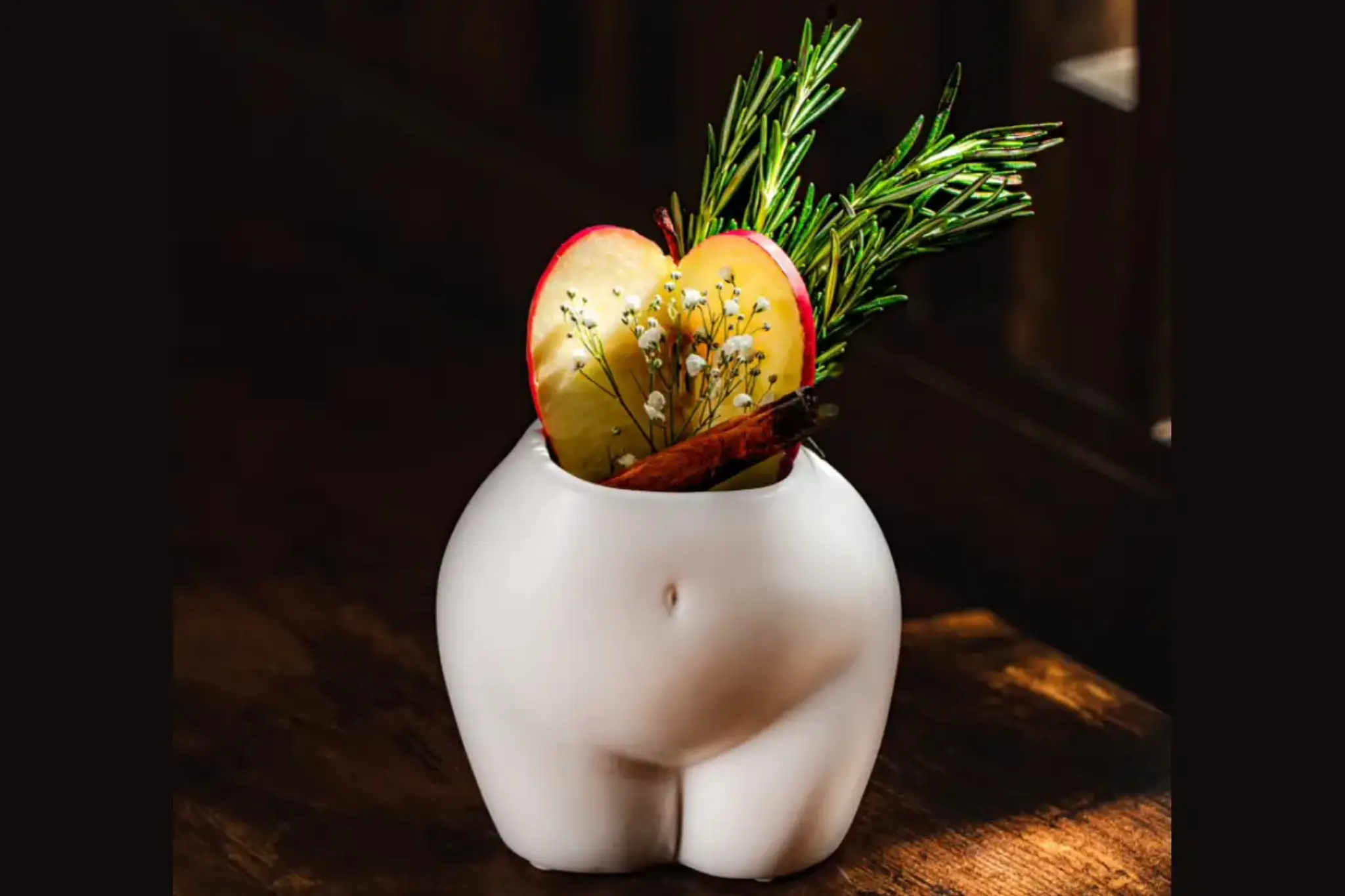 Executive Chef & Co-Founder webp A quirky white vase shaped like a buttocks, filled with apple slices, sprigs of rosemary, and a stick of cinnamon on a wooden table.