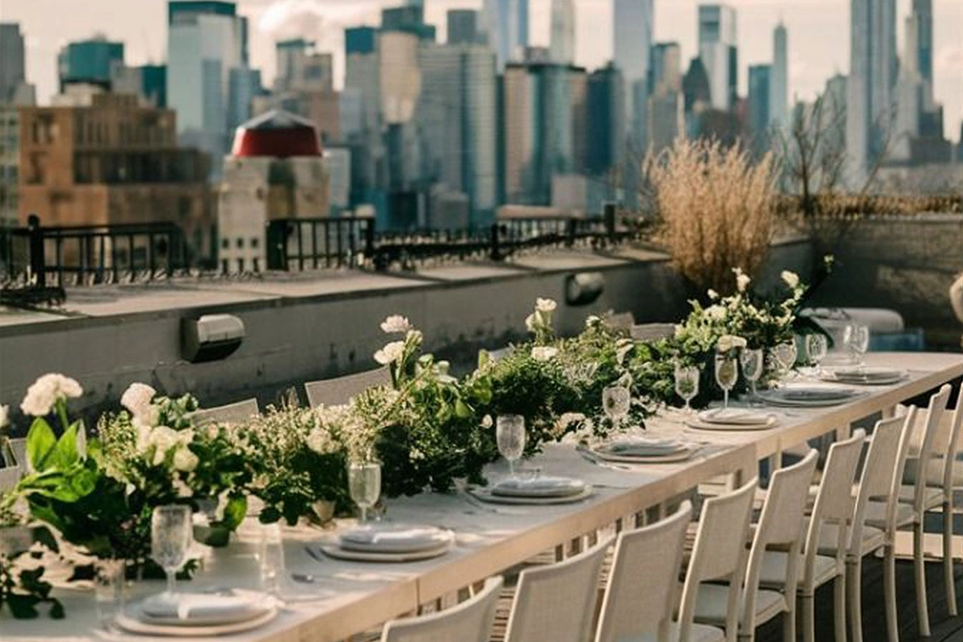 A beautifully arranged long dining table adorned with greenery and flowers, set against a stunning city skyline backdrop.